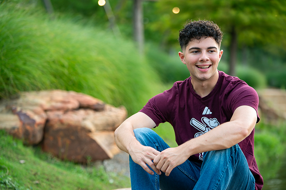 young man with curly black hair sitting outside in front of grass and rock background