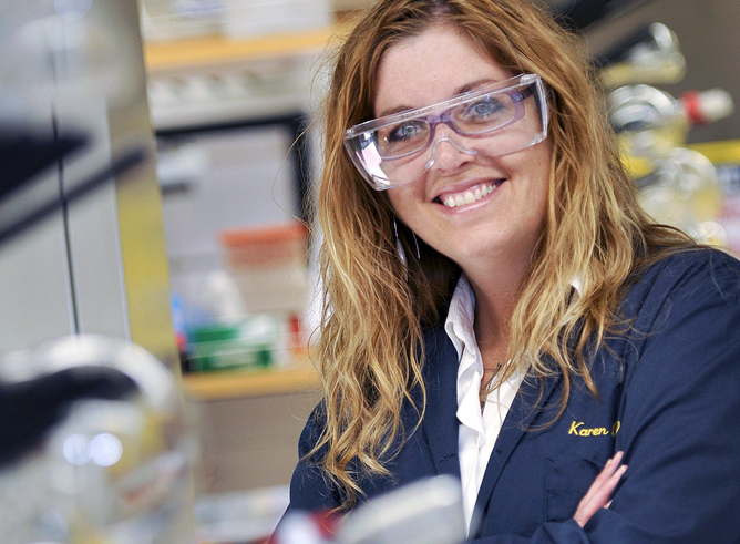 Texas A&M chemist Karen Wooley smiles for the camera in her laboratory while wearing goggles and her trademark blue labcoat