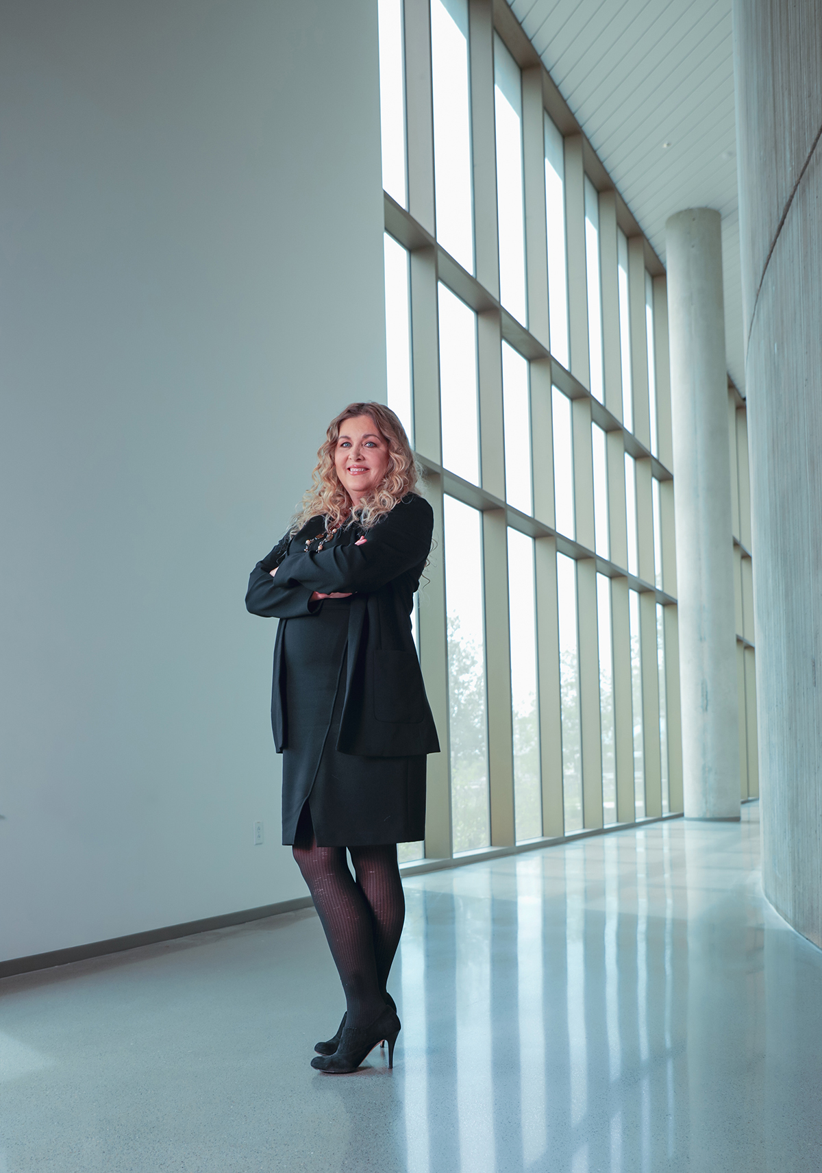 Texas A&M chemist Karen Wooley smiles for the camera, arms folded across her chest and standing in a hallway bathed in natural light from a bank of floor-to-ceiling windows