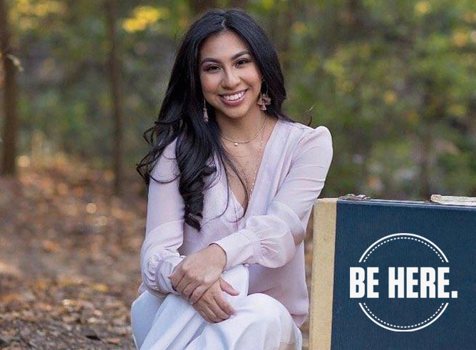 Texas A&M women's and gender studies major Odyssey Olmos smiles for the camera while sitting beside two suitcases in a tree-lined background setting