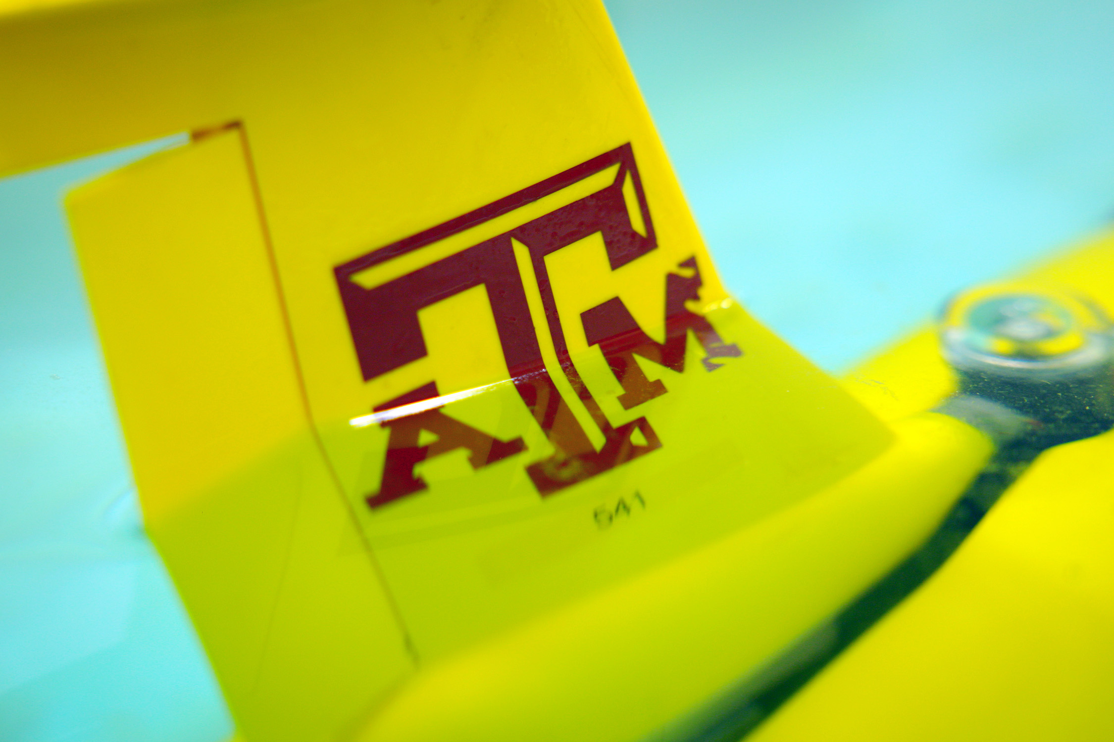 A yellow glider featuring a maroon Texas A&M University logo floats in a pool of water within a Geochemical and Environmental Research Group laboratory