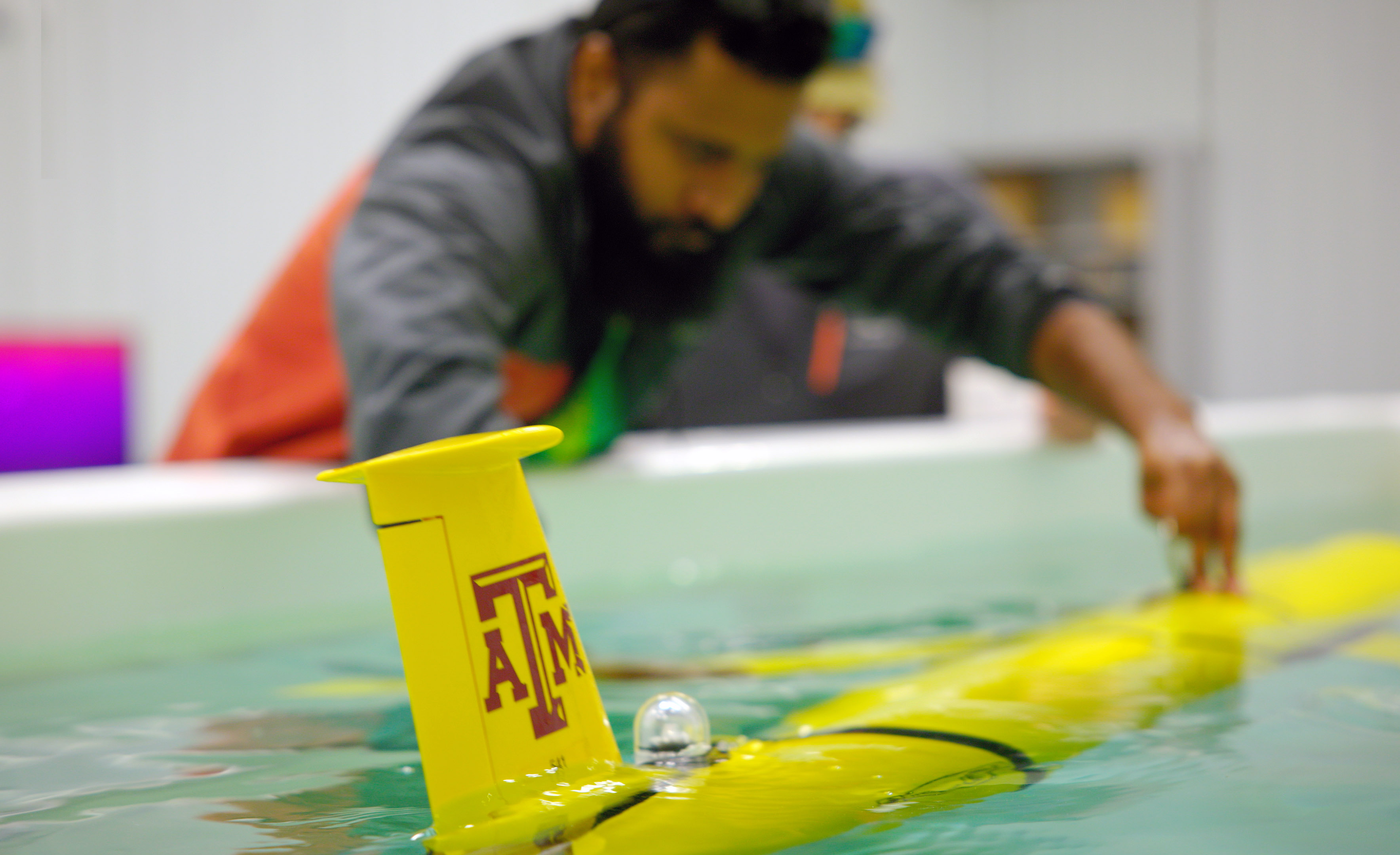 Postdoctoral researcher Sakib Mahmud observes a yellow glider featuring a maroon Texas A&M University logo as it floats in a pool of water within a Geochemical and Environmental Research Group laboratory