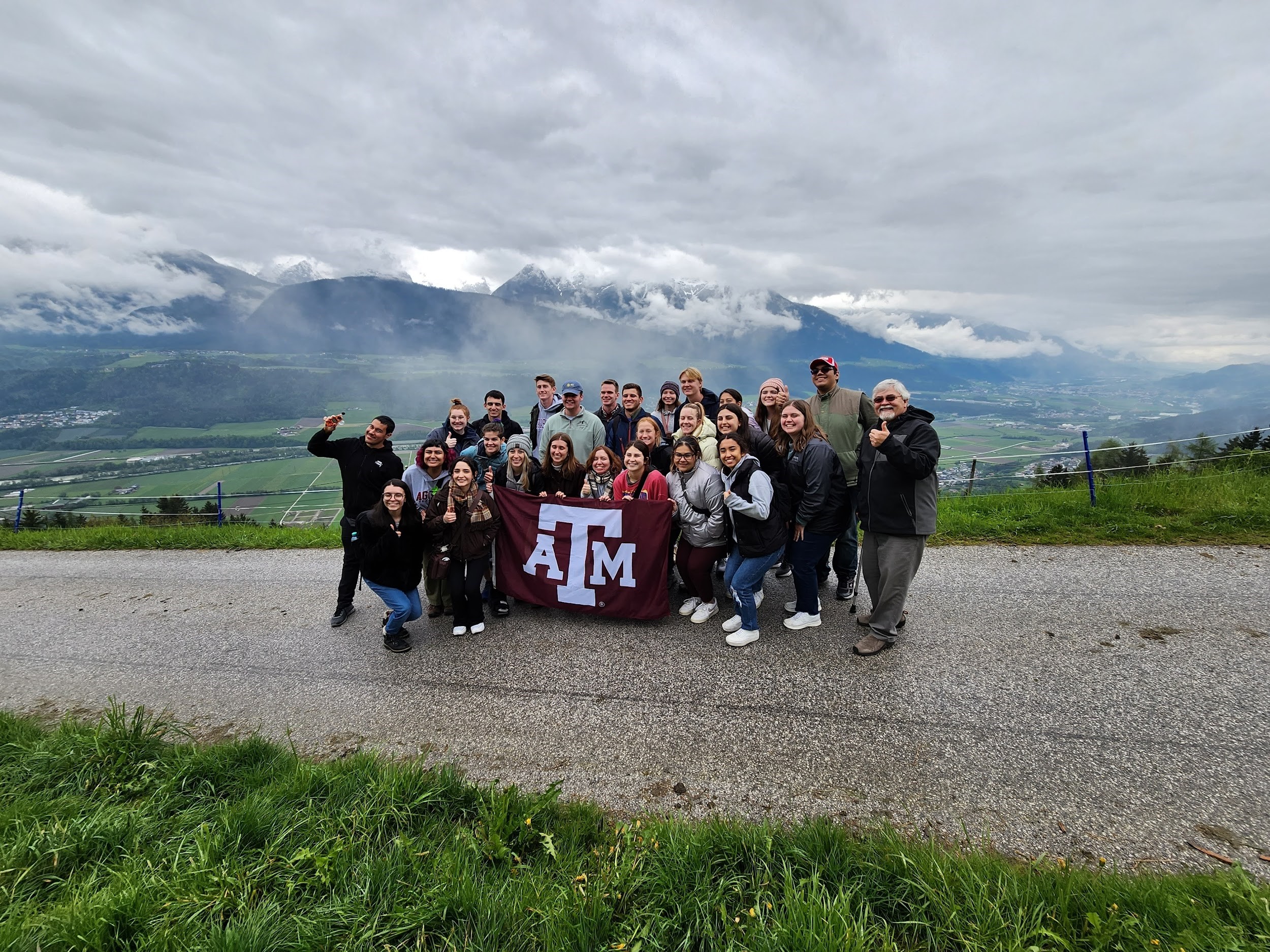 Texas A&M University students participating in the 2023 Student Experiences Abroad in Meteorology program pose with a maroon Texas A&M University flag with the Alps in the background