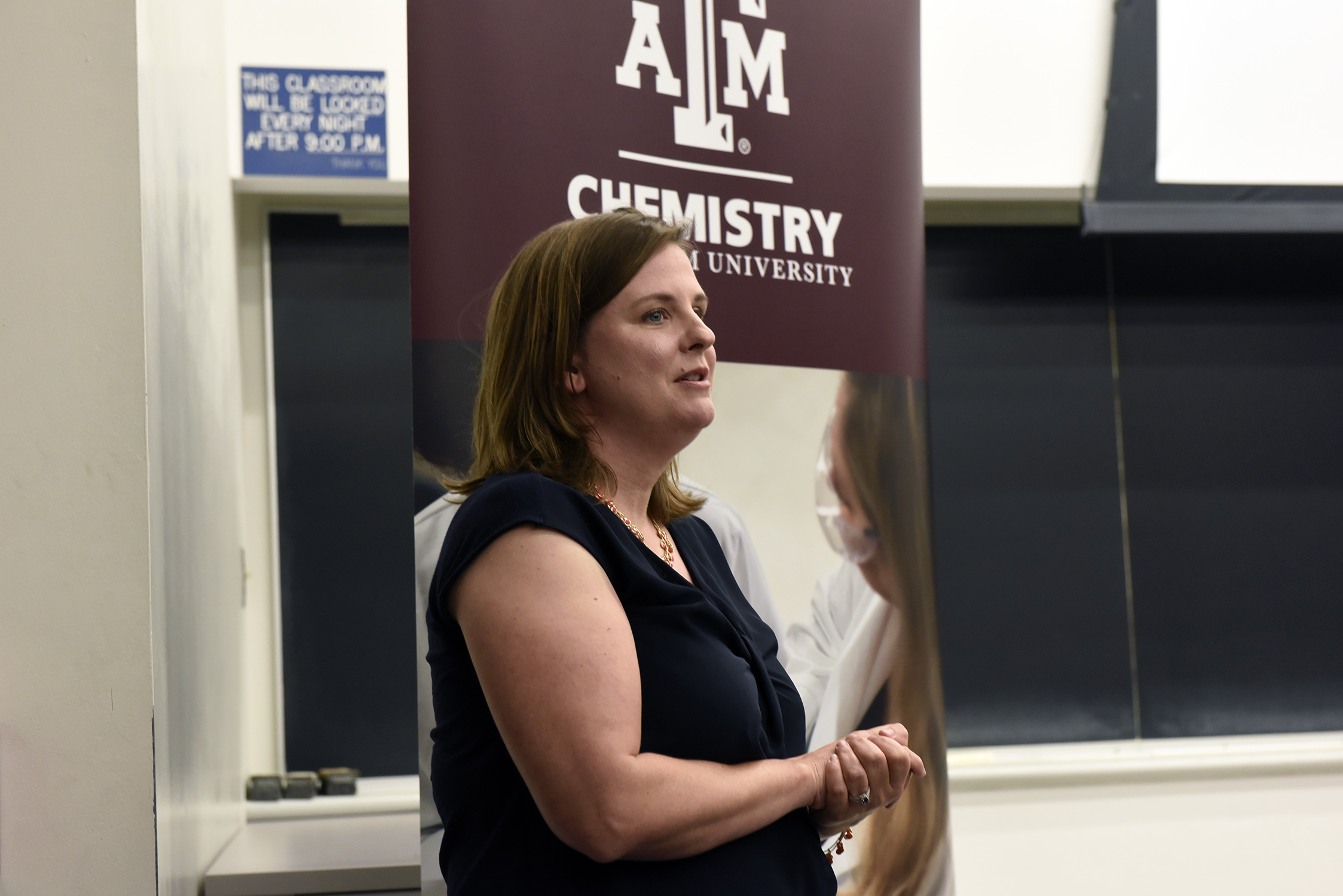 Texas A&M University chemist Joanna Goodey-Pellois speaks from the front of a classroom with a Texas A&M Chemistry banner and chalkboards in the background