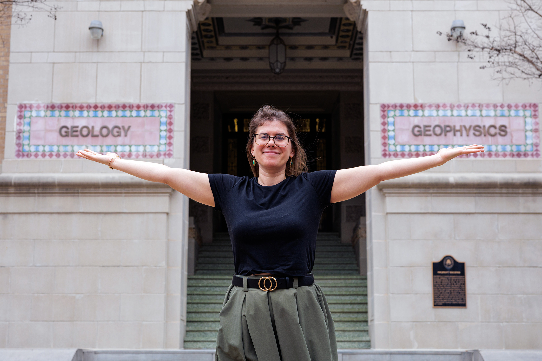 Texas A&M University Geology and Geophysics professor Brandi Lenz stands outside the Halbouty Building with outstretched arms, extending her palms in metaphorical support of the words "Geology" and "Geophysics" that appear in intricate tile format on the building's exterior wall