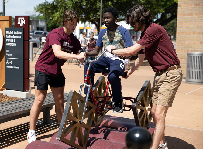 Two Texas A&M University student volunteers assist a male youth in riding a square-wheeled bicycle at the 2023 Physics and Engineering Festival, held April 1, 2023