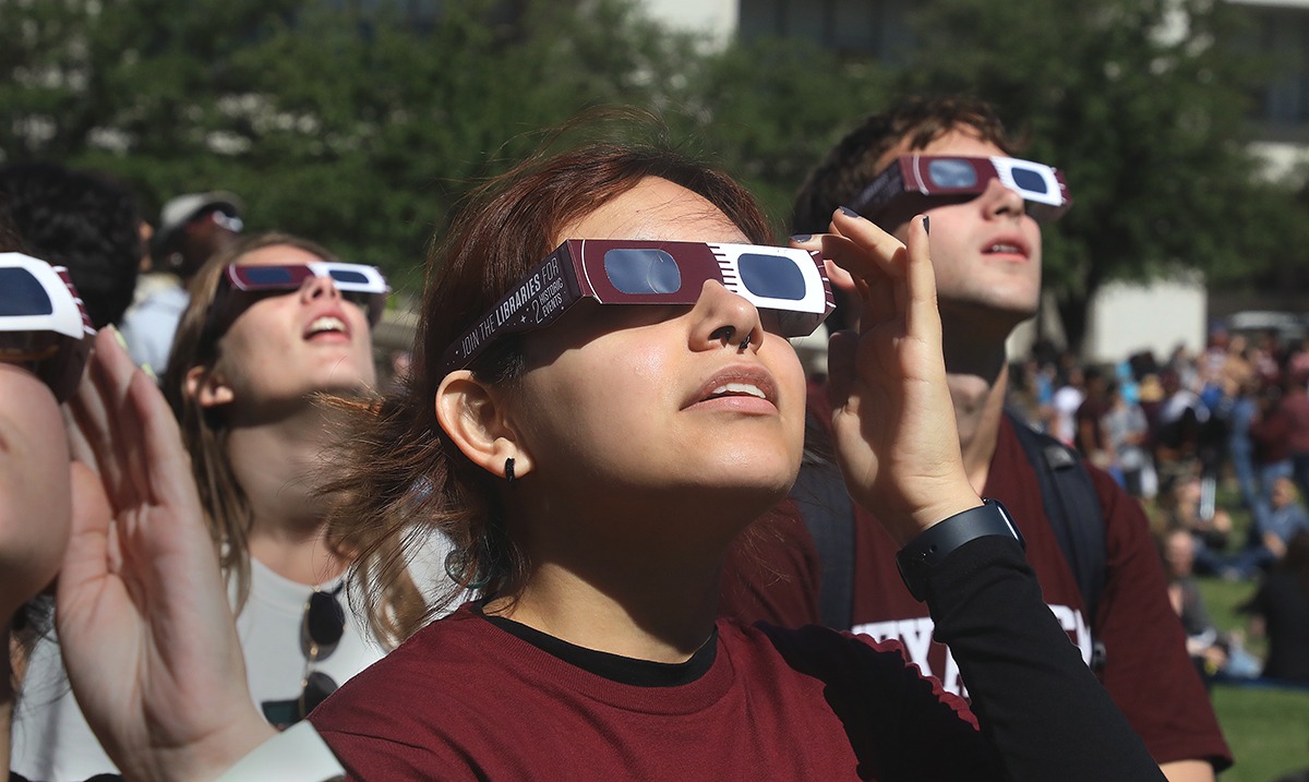 A group of Texas A&M University students look skyward while wearing Texas A&M-branded maroon and white eclipse glasses during the partial solar eclipse on October 14, 2023