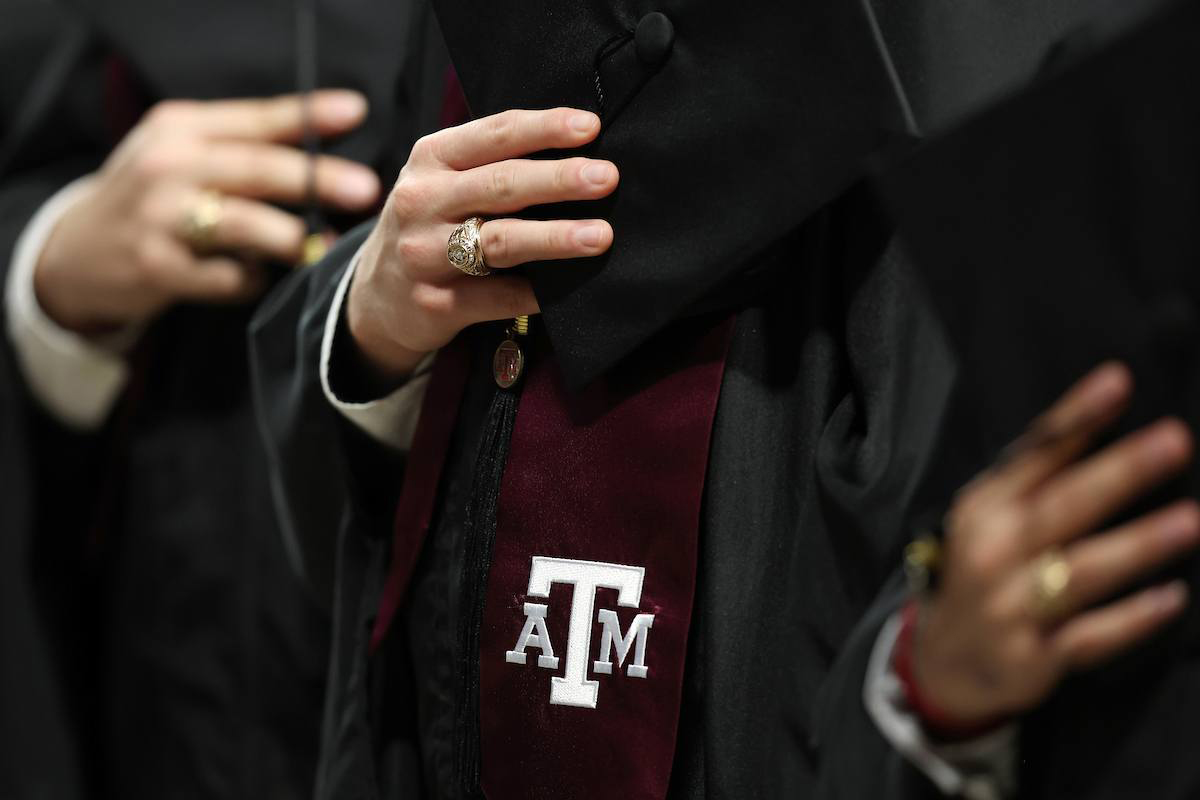 Close-up of Texas A&M University graduates in regalia, holding their mortar boards with their Aggie Rings and maroon stoles featuring the white aTm logo visible