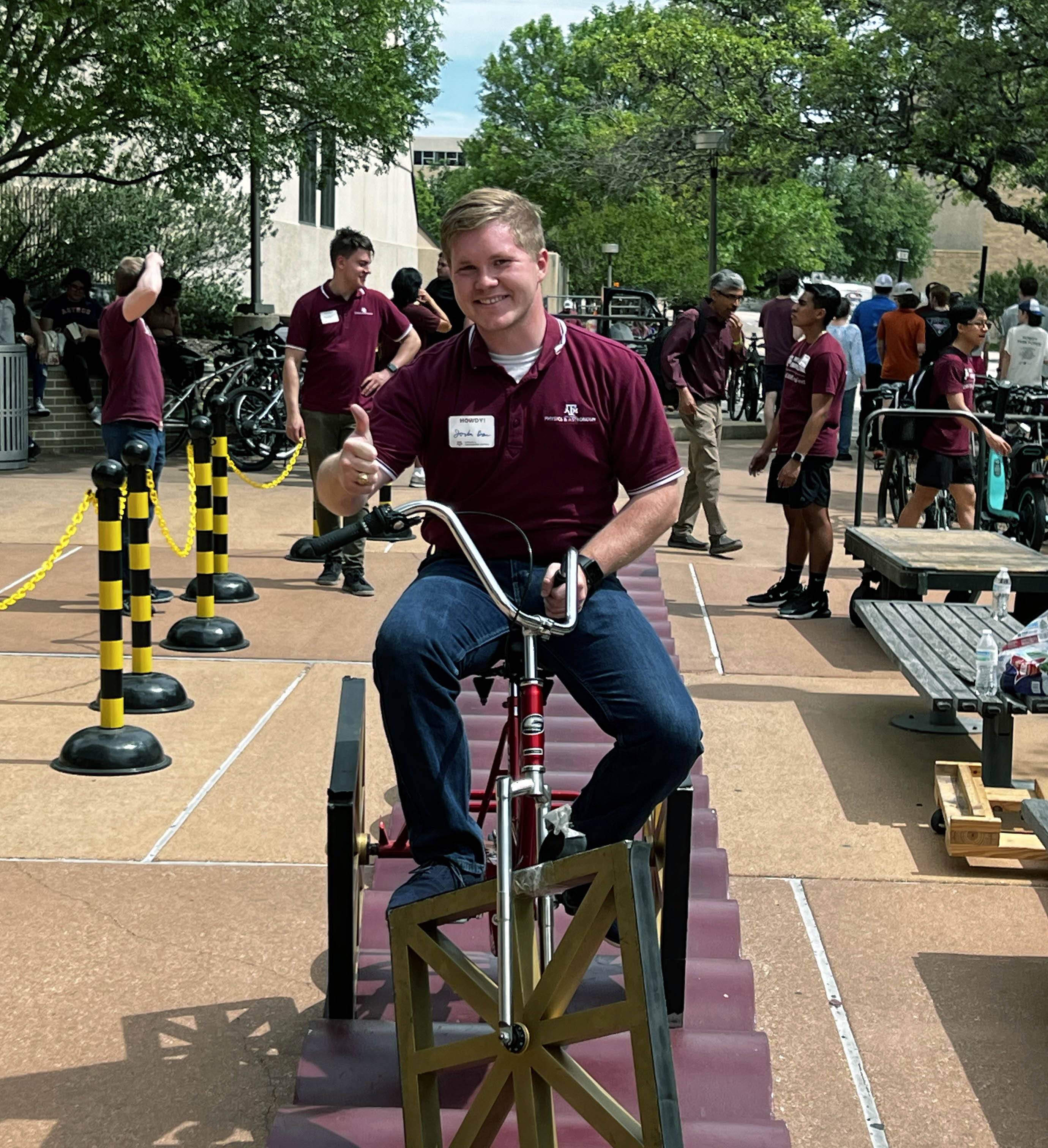 2024 Texas A&M University physics graduate Jordan Bass flashes a gig 'em while riding the square-wheeled bicycle during the 2024 Physics and Engineering Festival on the Texas A&M campus
