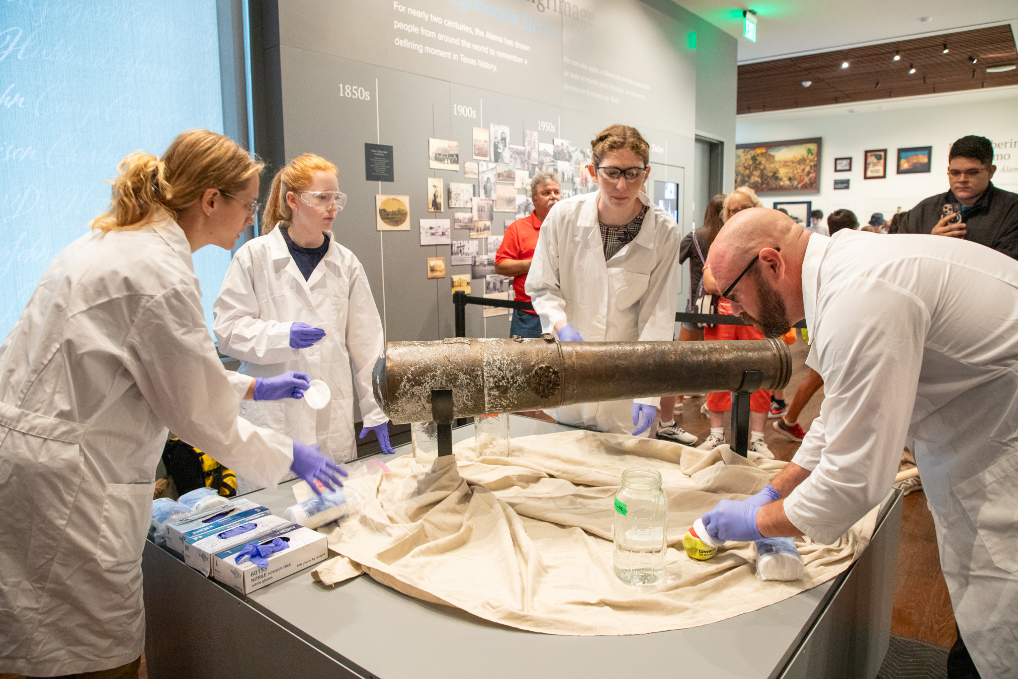 Three female graduate students from Texas A&M University wearing white lab coats, goggles and blue latex gloves work with Texas A&M anthropologist Chris Dostal to clean a bronze cannon at the Alamo Museum in San Antonio