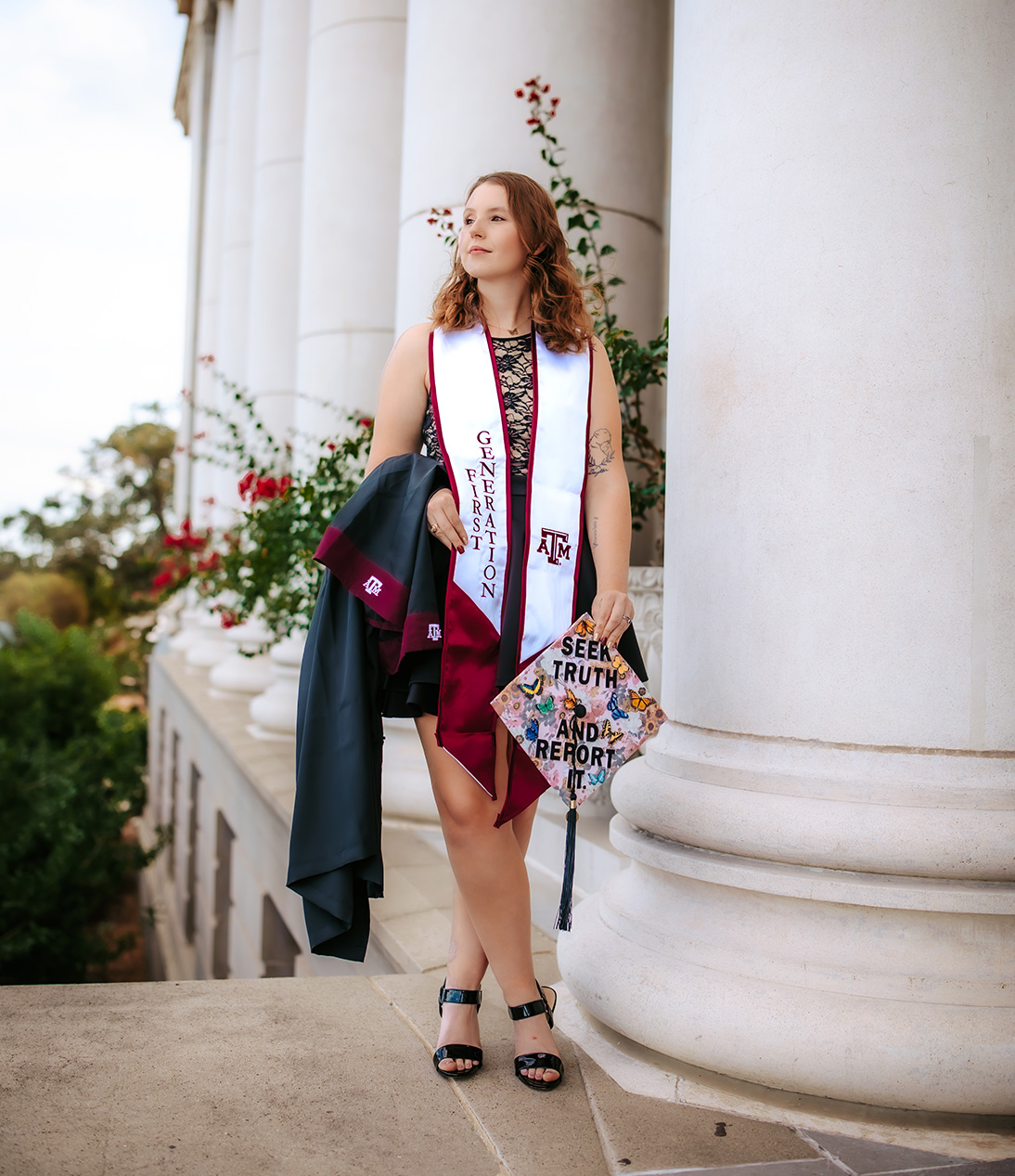 Megan standing confidently in front of the large white columns of the Jack K. Williams Administration Building, wearing a "First Generation" stole, holding a decorated graduation cap that reads "Seek Truth and Report It," and carrying her Texas A&M gown over her arm.