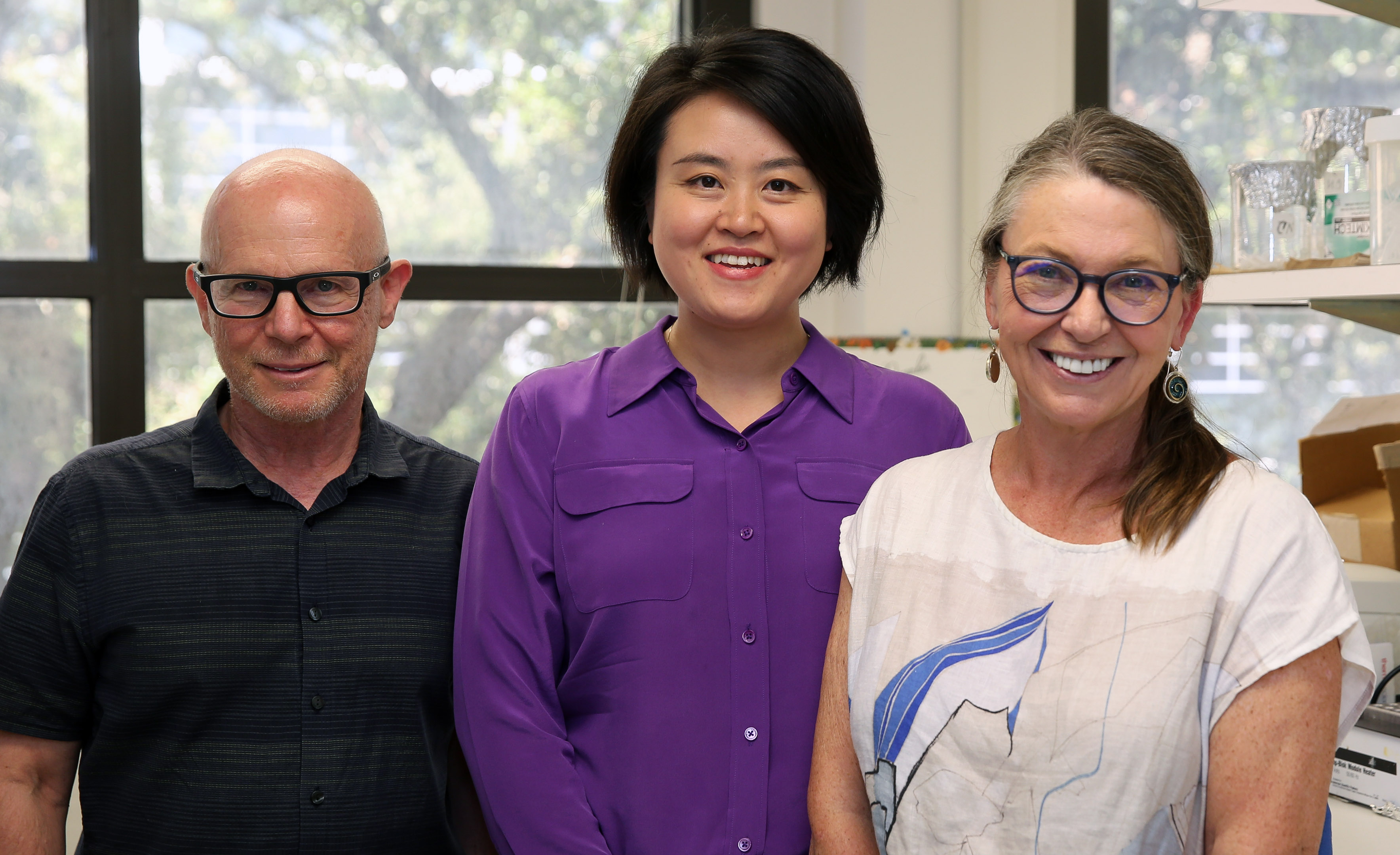 Texas A&M University biologists Dr. Matthew Sachs, Dr. Wanhe Li and Dr. Deborah Bell-Pedersen are smiling in an office setting, with Dr. Li in a purple shirt flanked by two others, Dr. Sachs wearing glasses and a black shirt, and Dr. Bell-Pedersen in a white shirt with a blue abstract design.