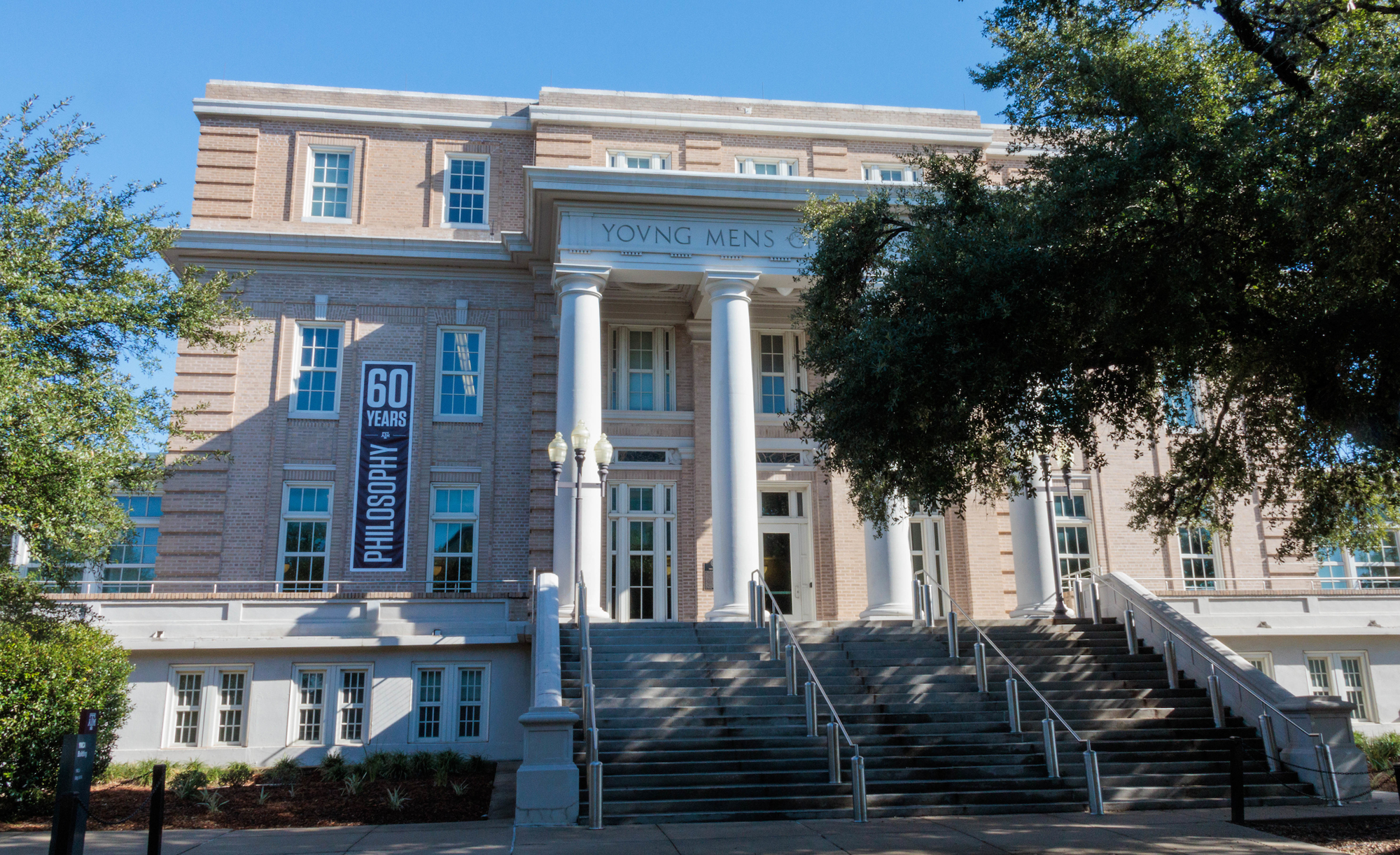 Exterior view of the Young Men’s Christian Association building on the campus of Texas A&M with a banner celebrating 60 years of philosophy on the left side of the building.