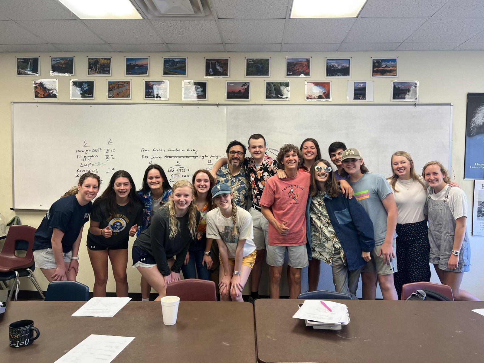 Group of students and a teacher smiling in a classroom with posters and a whiteboard in the background.