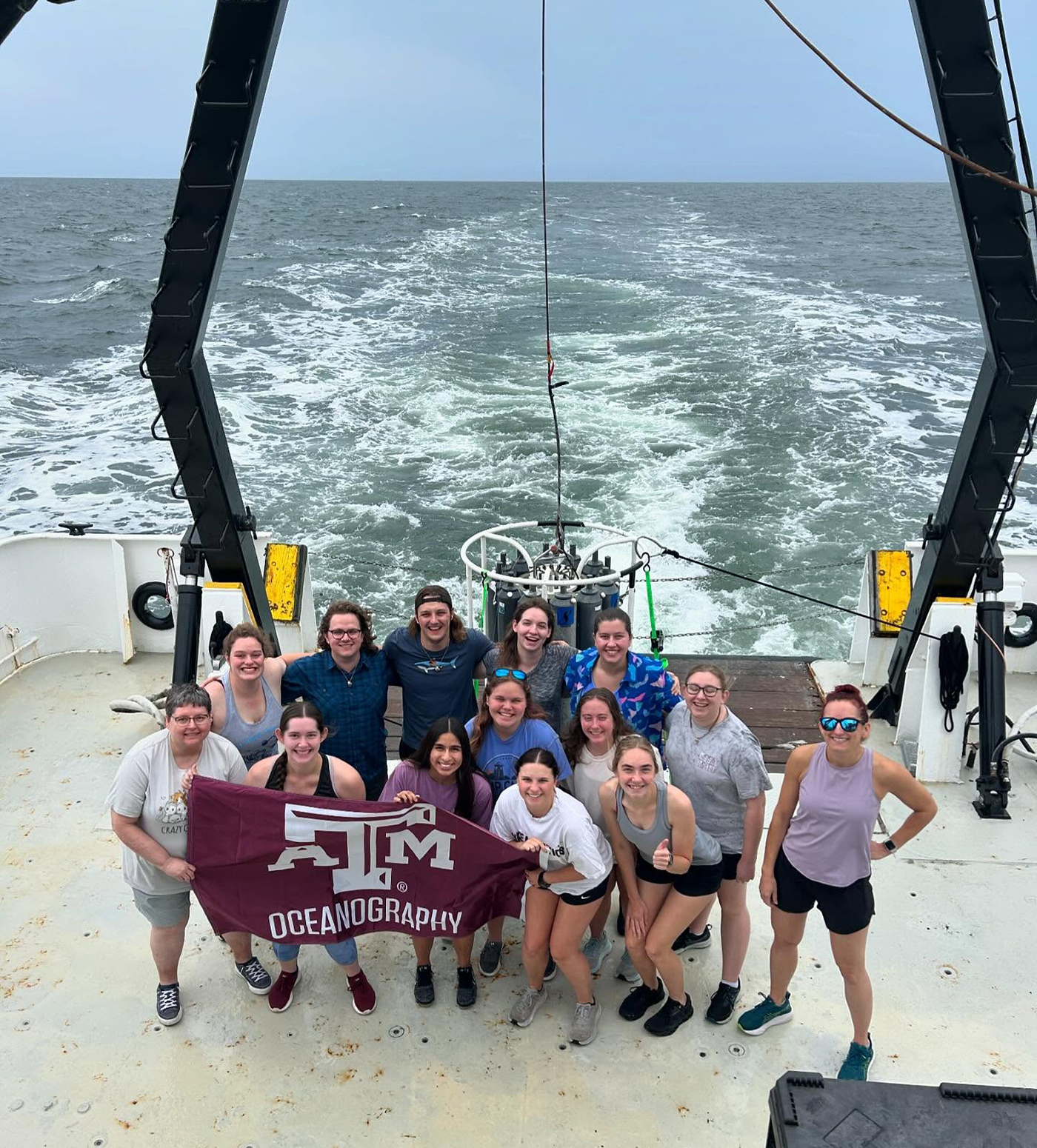 Group of students and faculty holding a Texas A&M Oceanography banner on the deck of a boat with the ocean in the background.