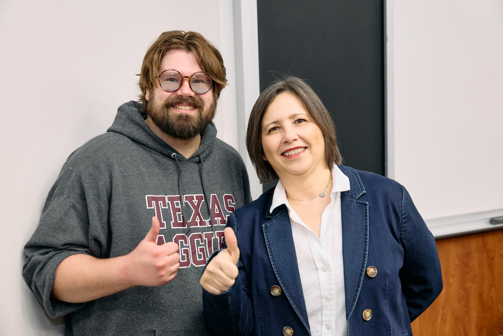 Jeremy in a Texas A&M sweatshirt and Oksana in a navy blazer, standing in a classroom with their thumbs up.