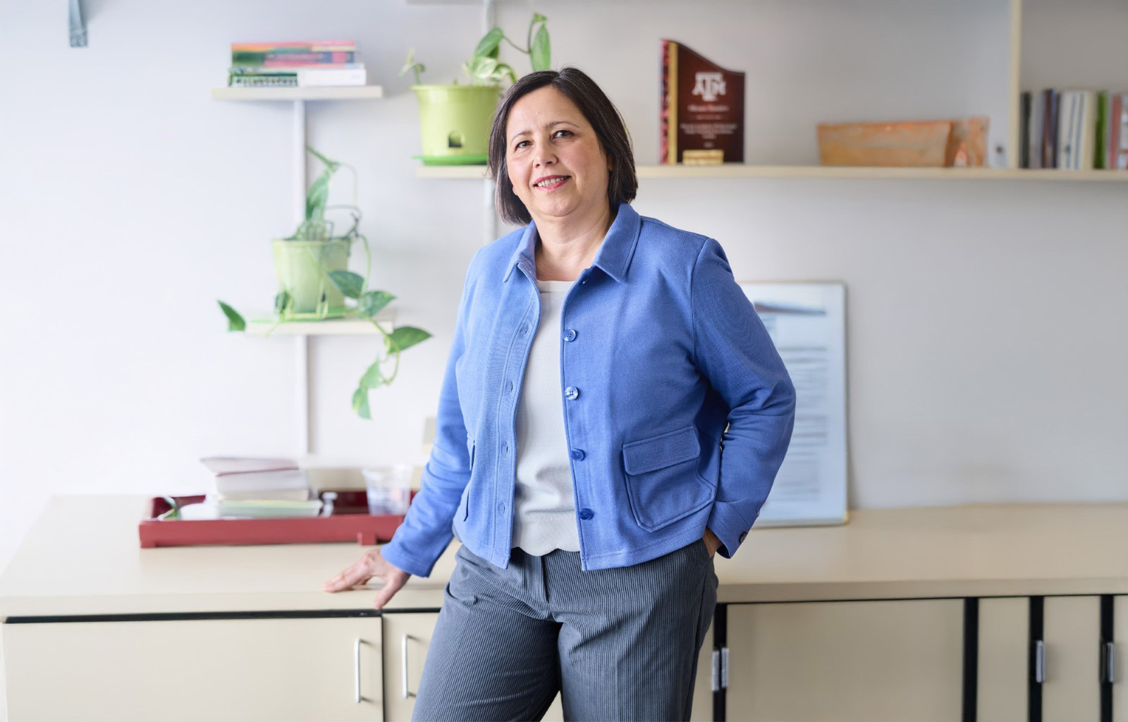 Dr. Oksana Shatalov in business attire stands confidently in an office setting, with a desk and books in the background.