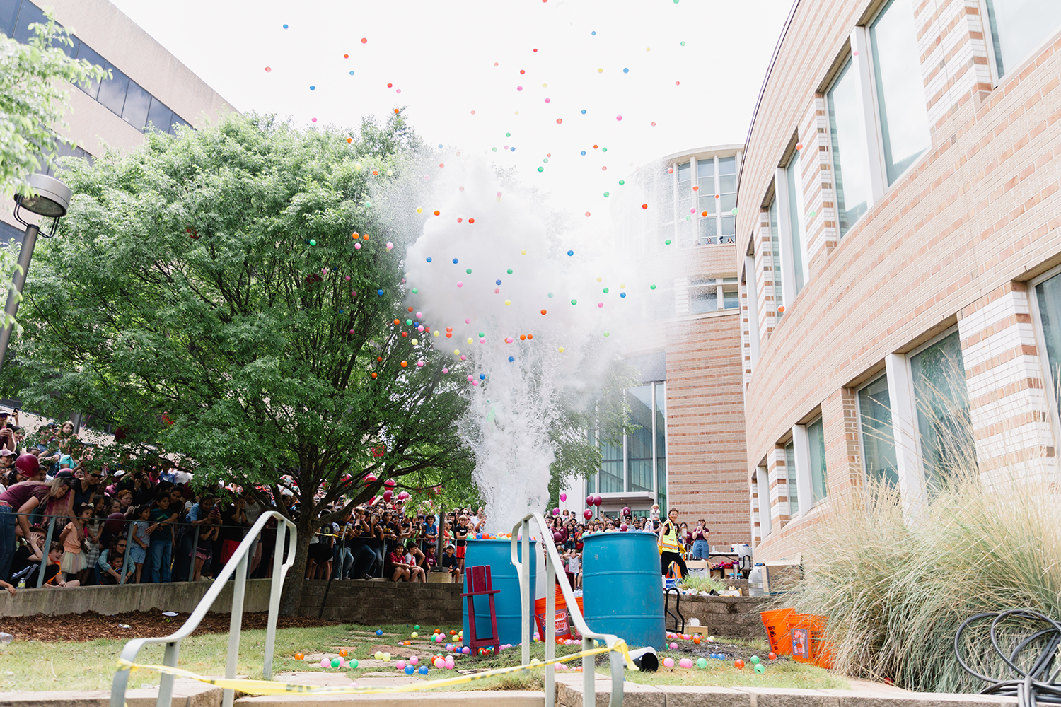 A lively 2023 Physics Festival at Texas A&M University, where a crowd is gathered with balloons emerging explosively from blue barrels amidst a campus setting.