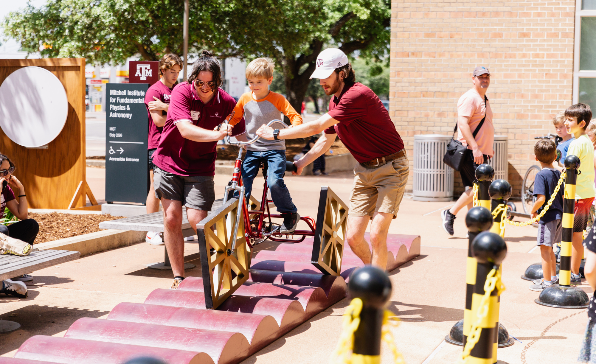Two adults assist a child riding a bicycle with squae wheels on an elevated, wavy track at the Texas A&M 2023 Physics Festival, with other spectators in the background.
