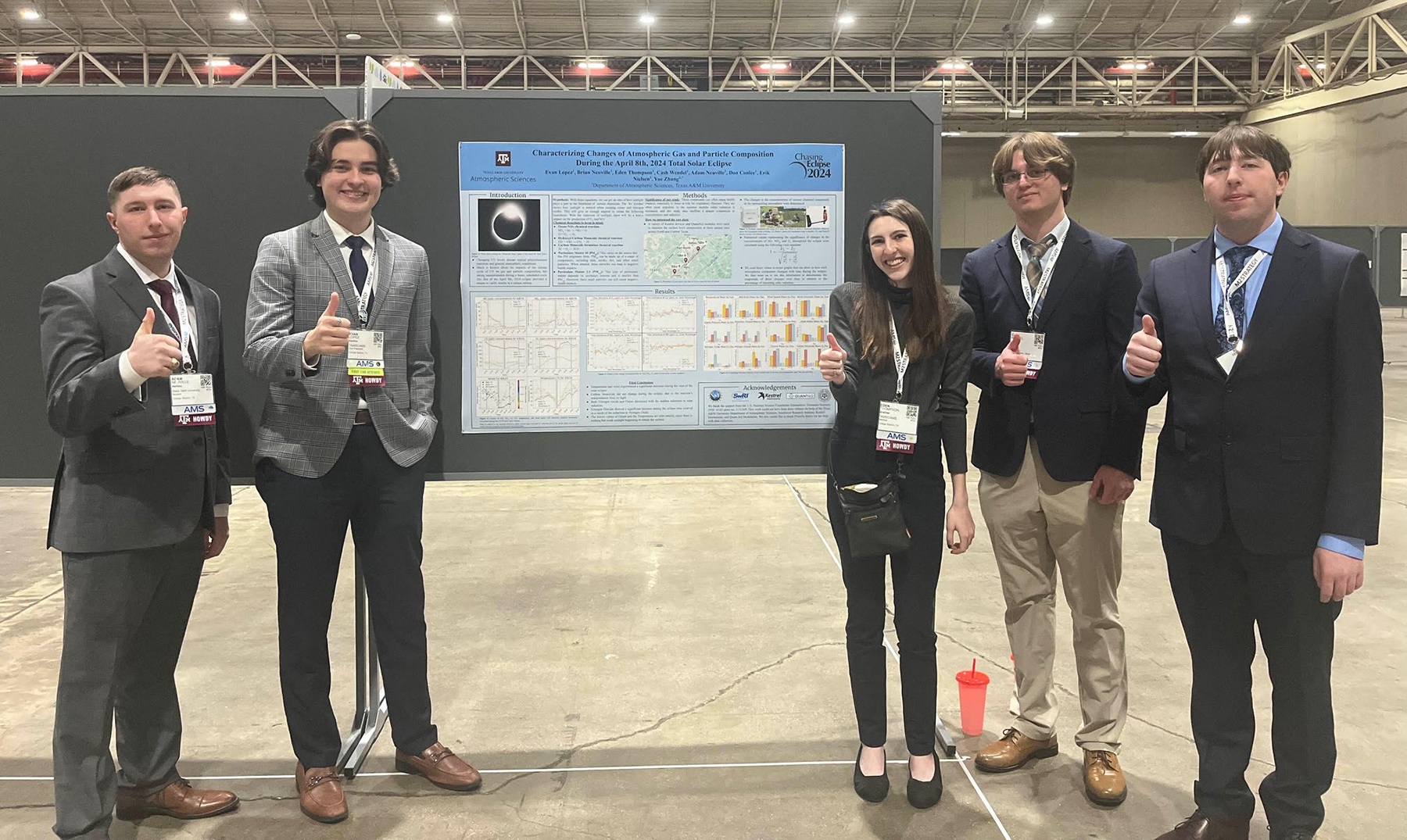 Five individuals stand confidently in front of a scientific poster at a conference, each wearing a name badge and formal attire. They are posing with thumbs up gestures.