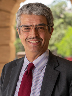 Portrait of a smiling individual wearing glasses, a dark suit, a white shirt, and a red tie, standing in front of a blurred architectural background.