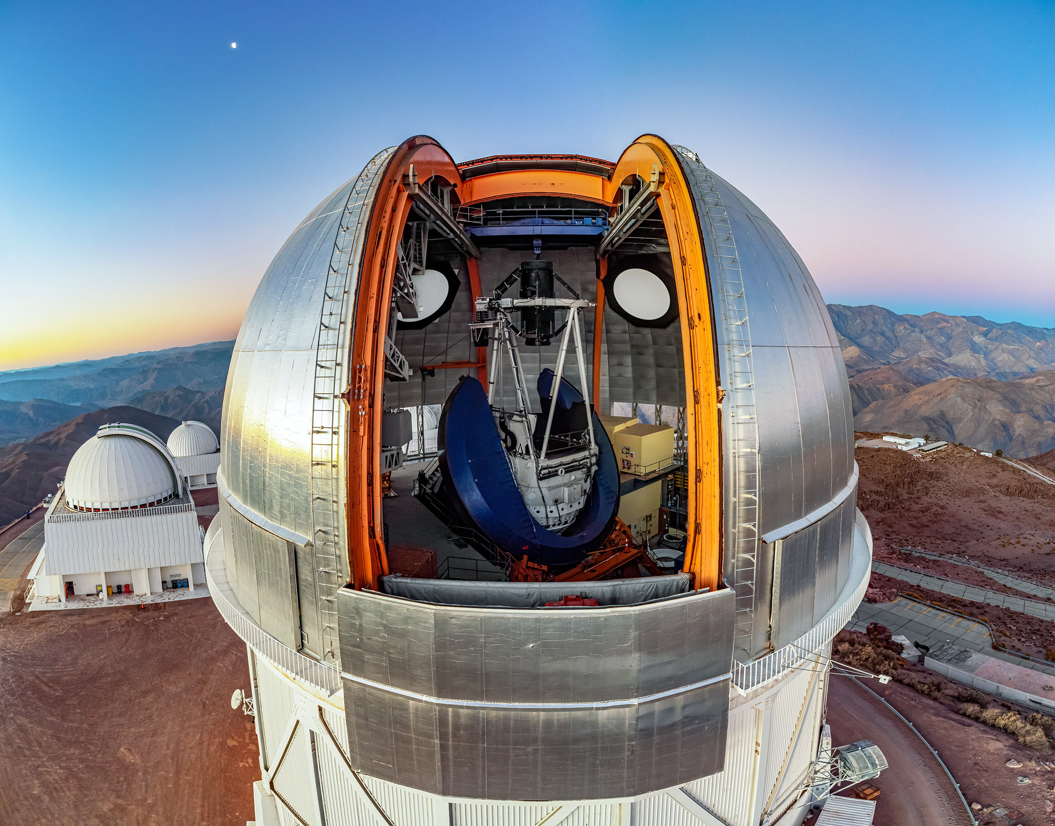 Aerial drone image of the Victor M. Blanco Telescope at Cerro Tololo Inter-American Observatory in Chile against a backdrop of twilight skies featuring the evening star otherwise known as the planet Venus, with the Andes Mountains and both the SMARTS 1.5-meter and 0.9-meter telescopes in the background.