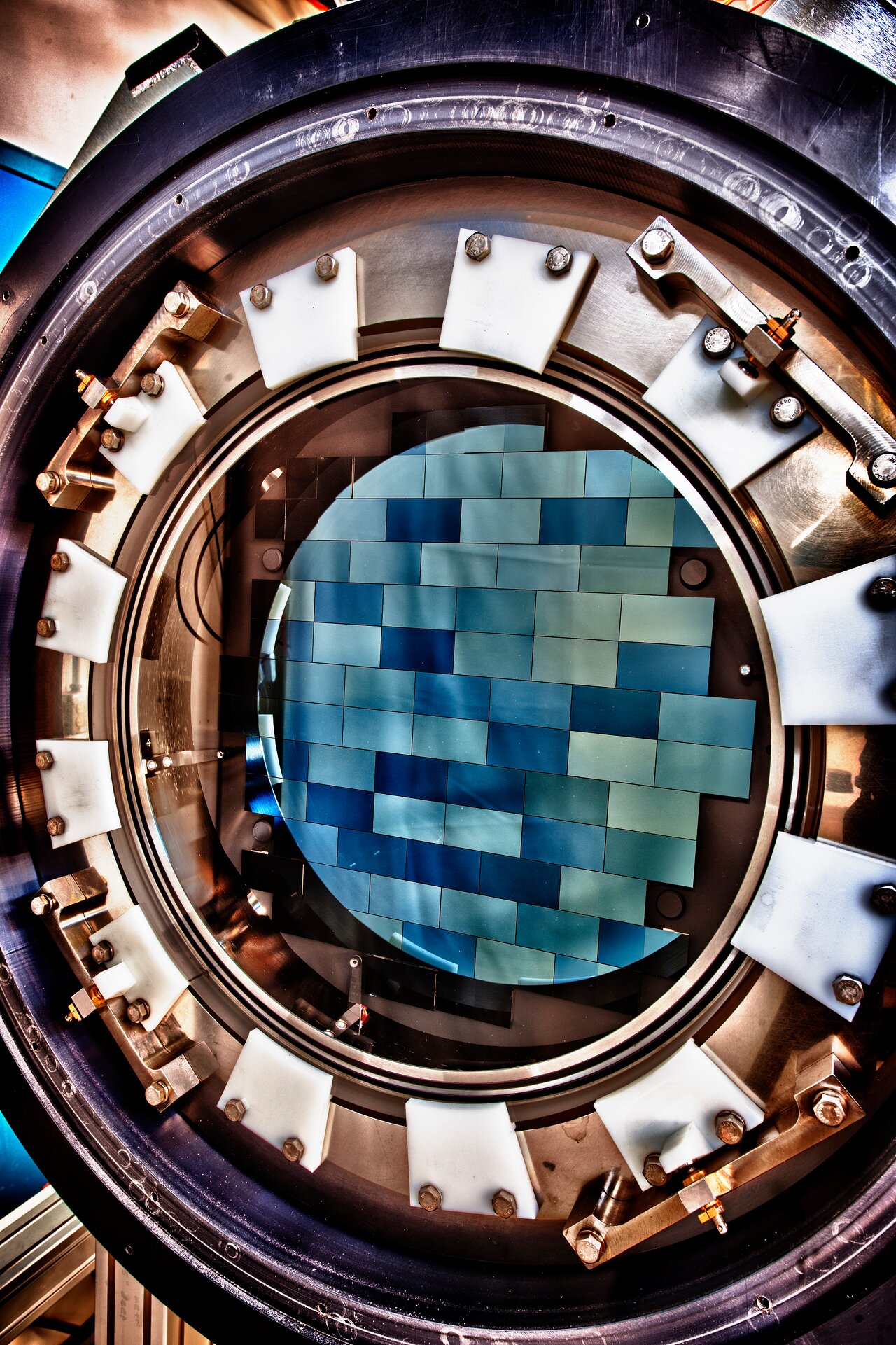 Close-up image of the Dark Energy Camera, a circular mechanical assembly featuring a mosaic of blue tiles – its 62 charge-coupled devices – at its center surrounded by bolts and metal edging.
