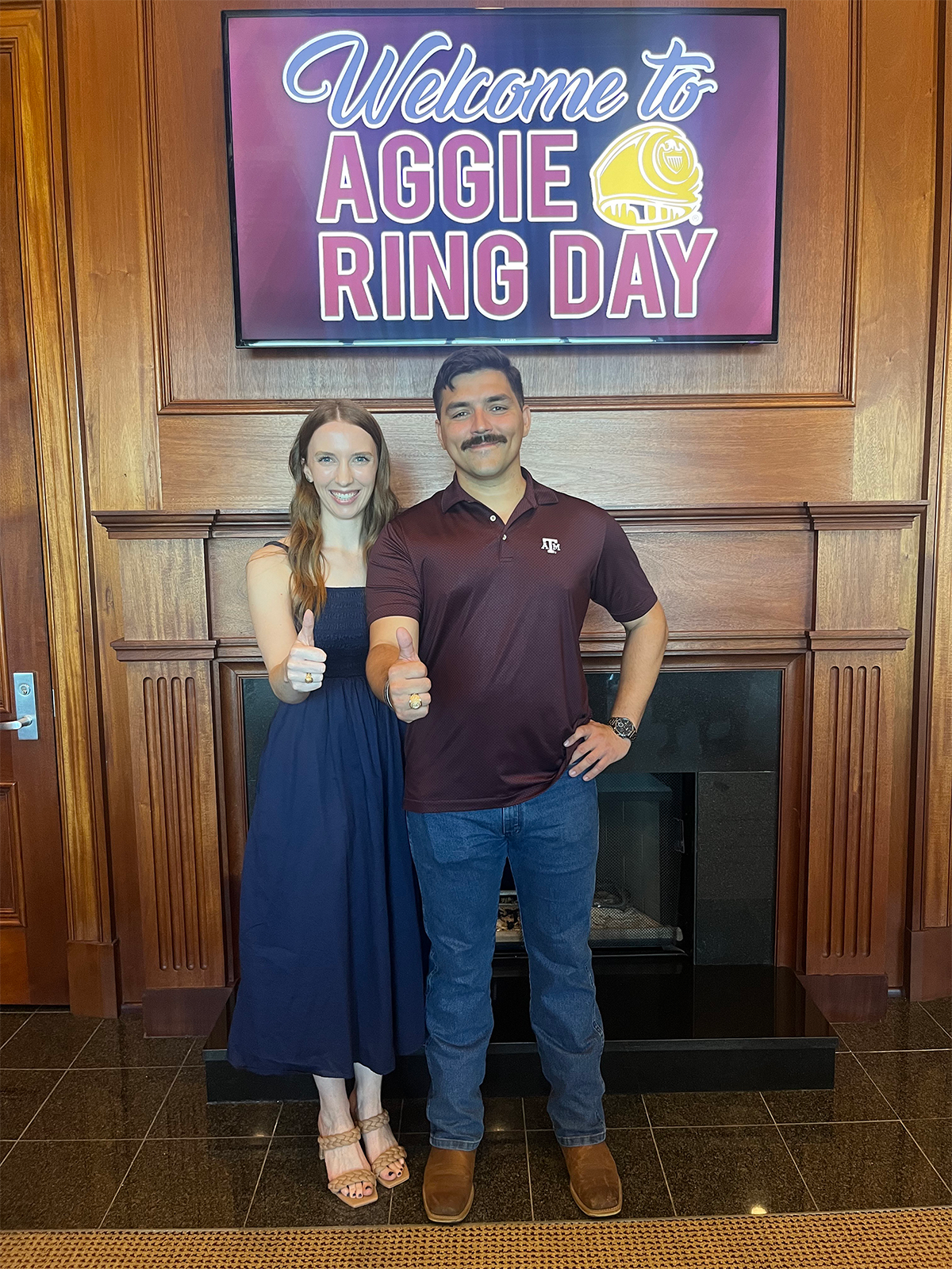 Two individuals standing in front of a "Welcome to Aggie Ring Day" banner, each holding up a ring towards the camera.