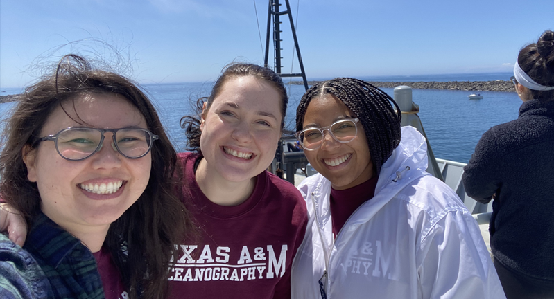 Dr. Sarah Hu, Alexis Adams and Kayla Nedd s smiling for a selfie on a sunny day by the ocean, with all three wearing Texas A&M University apparel.