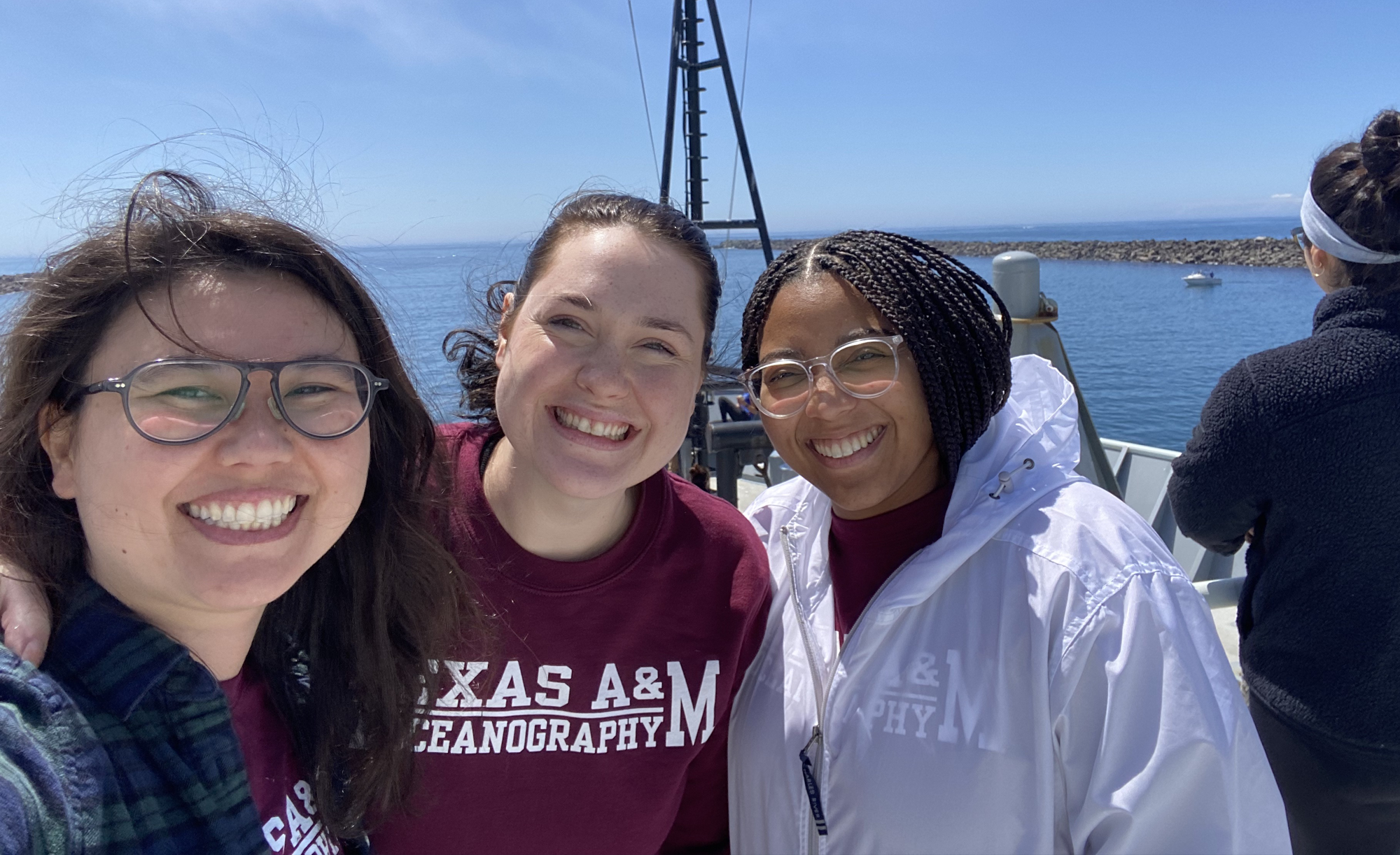 Dr. Sarah Hu, Alexis Adams and Kayla Nedd s smiling for a selfie on a sunny day by the ocean, with all three wearing Texas A&M University apparel.