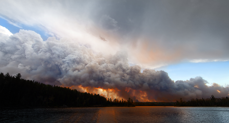 Image showing a large wildfire with thick smoke clouds above a forest near a lake under a dramatic sky.