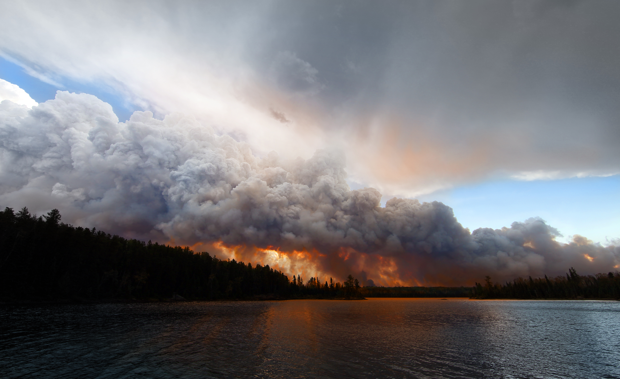 Image showing a large wildfire with thick smoke clouds above a forest near a lake under a dramatic sky.