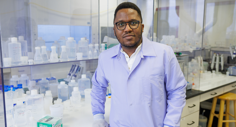 Dr. Lucien Nana Yobo in a lab coat standing in a laboratory filled with chemical bottles and lab equipment.