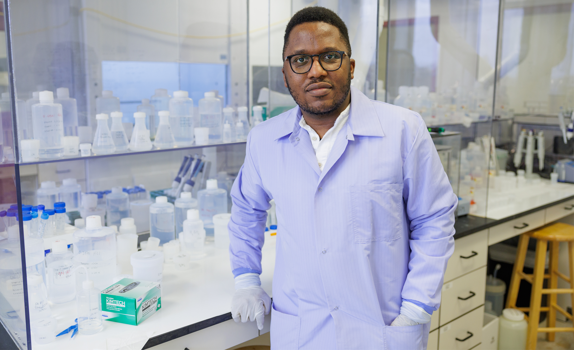 Dr. Lucien Nana Yobo in a lab coat standing in a laboratory filled with chemical bottles and lab equipment.