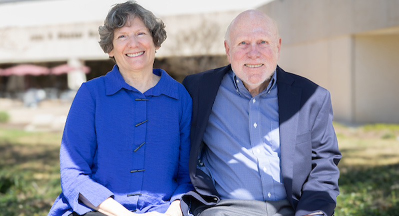 Two smiling individuals sitting closely together outside, with a modern building in the background. The person on the left is wearing a blue shirt, and the person on the right is in a light blue shirt.
