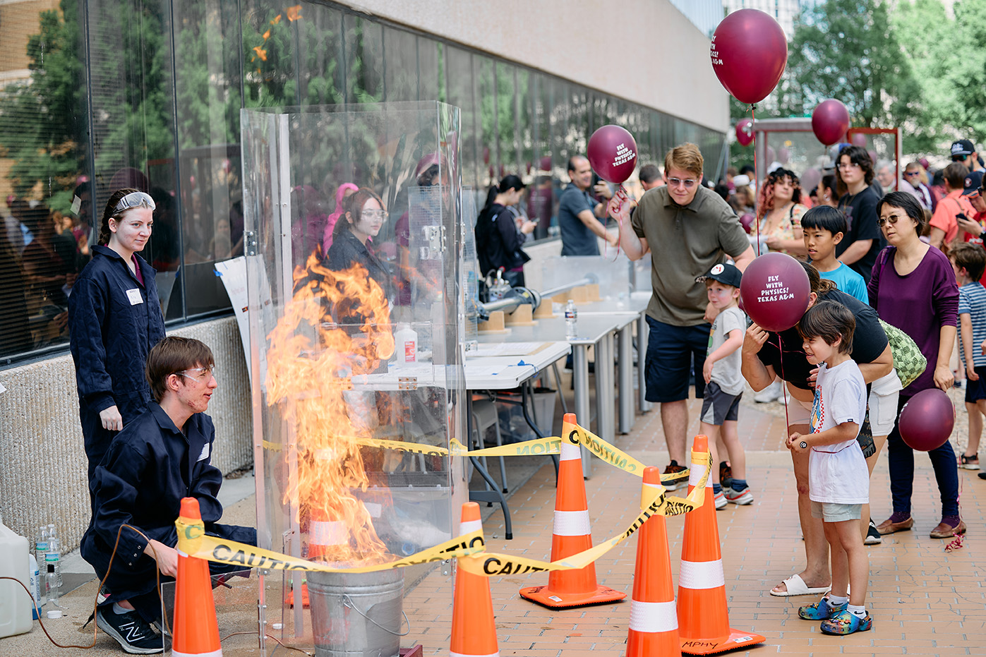 : Festival attendees look on with awe as two students demonstrate a physics experiment with fire. The students are in PPE and the onlookers are holding maroon balloons that say “Fly with Physics! Texas A&M.”