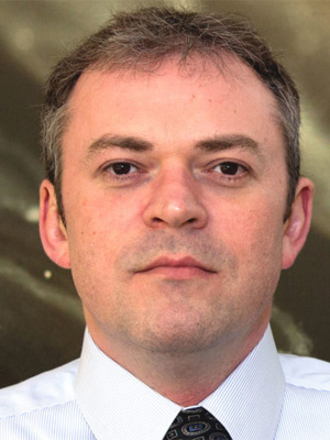 Portrait of Safonov, wearing a white shirt and patterned tie, with a focused expression.