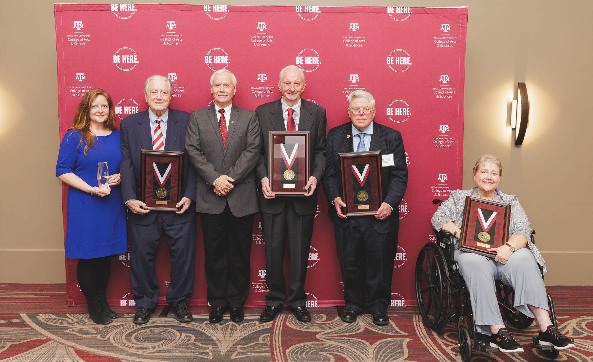 Group of six individuals holding awards, standing in front of a Texas A&M University banner with the slogan "Be Here.