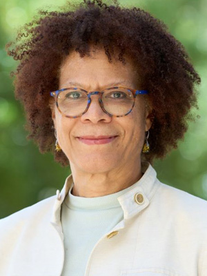 Portrait of a smiling individual with curly hair and glasses, wearing a light jacket over a turquoise top.