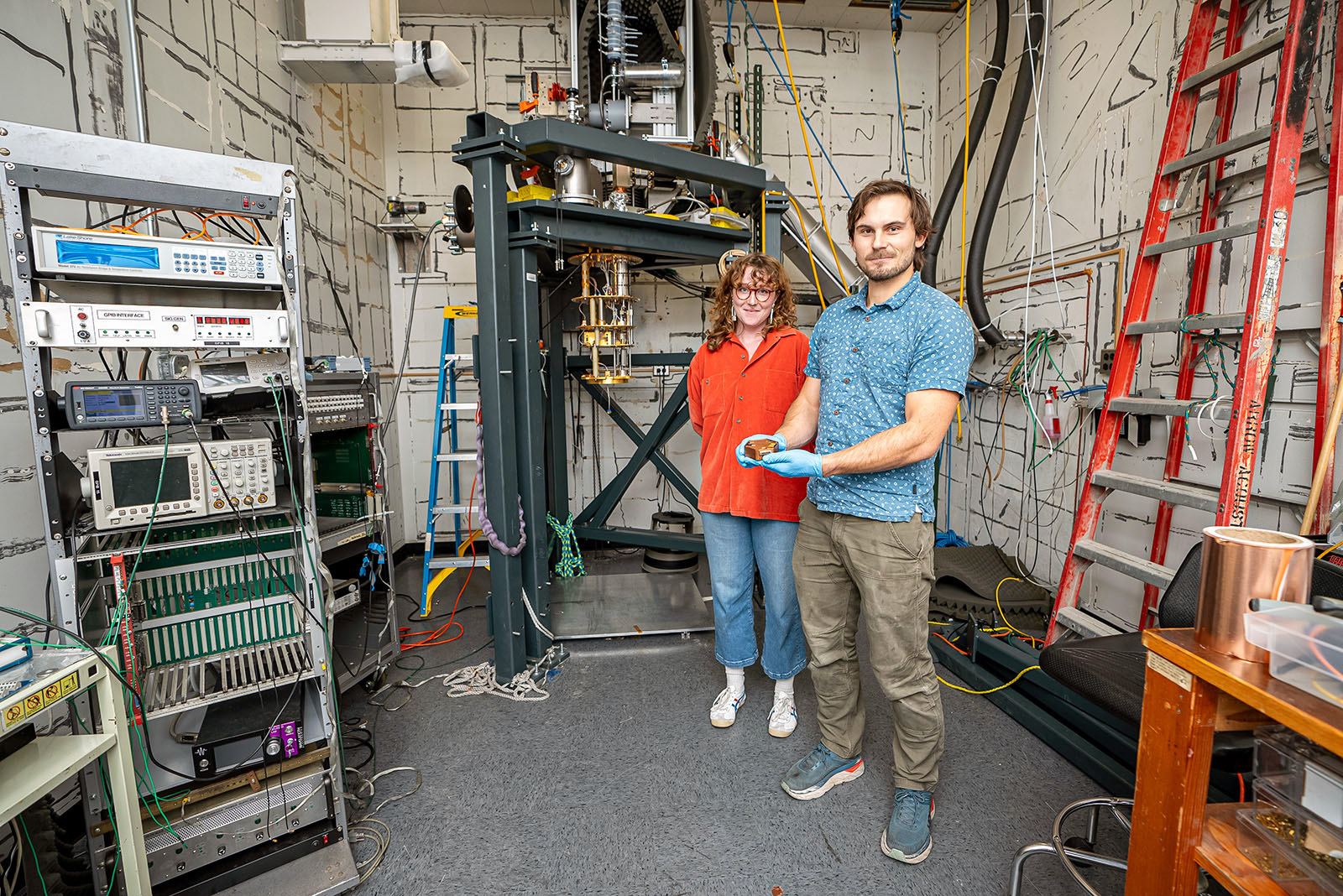 Two people stand in a lab room with a rack of electronics and a metal frame suspending equipment. One person holds a small hexagonal detector.