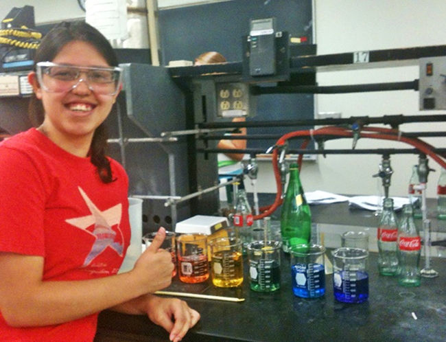 Samantha Yruegas in a red T-shirt with a "Texas" logo stands smiling in a laboratory, surrounded by various colored liquids in glassware and several Coca-Cola bottles.