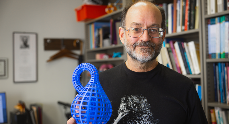 Person holding a 3D-printed blue object, wearing a T-shirt with a raven illustration, standing in front of a bookshelf.