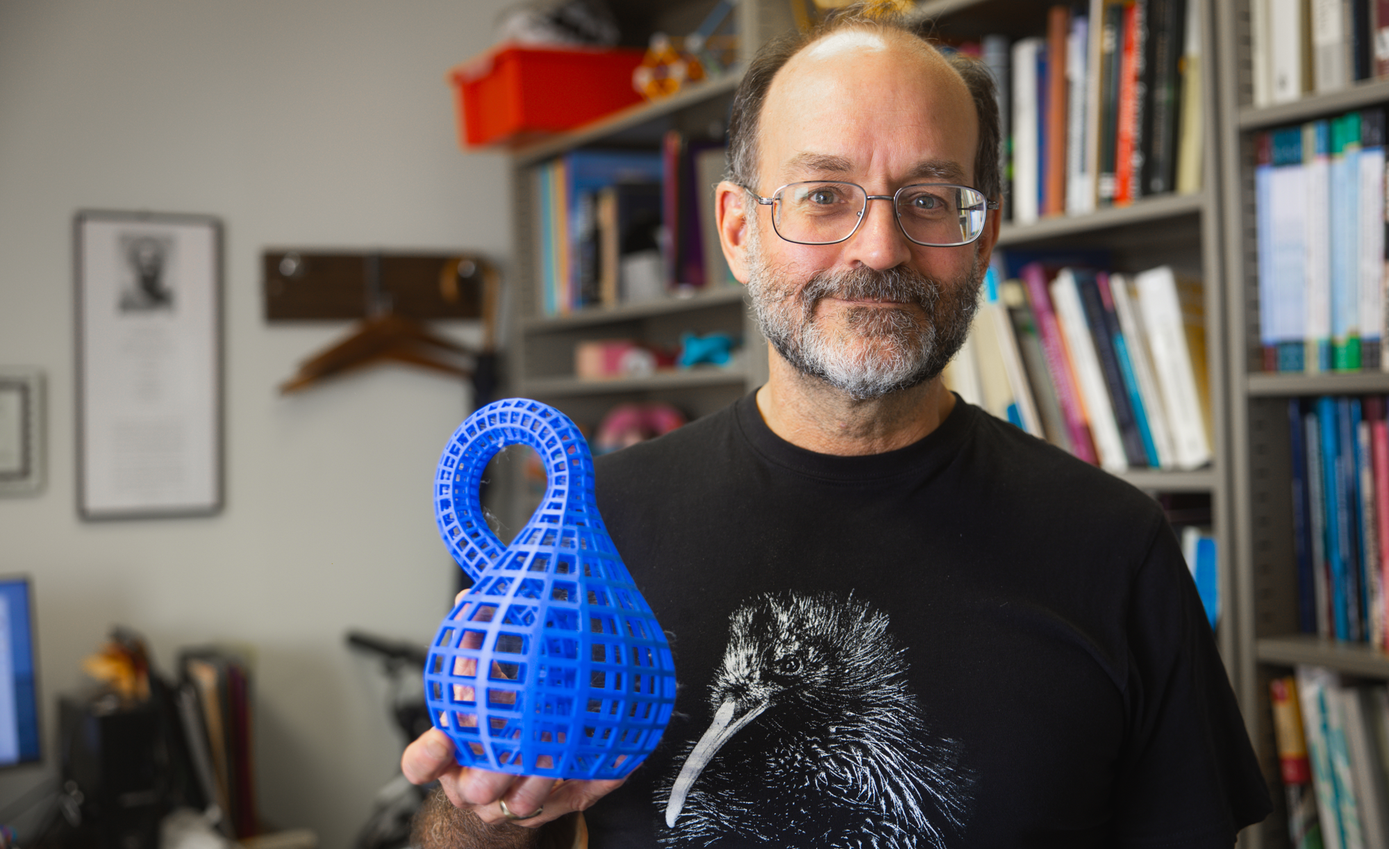 Person holding a 3D-printed blue object, wearing a T-shirt with a raven illustration, standing in front of a bookshelf.