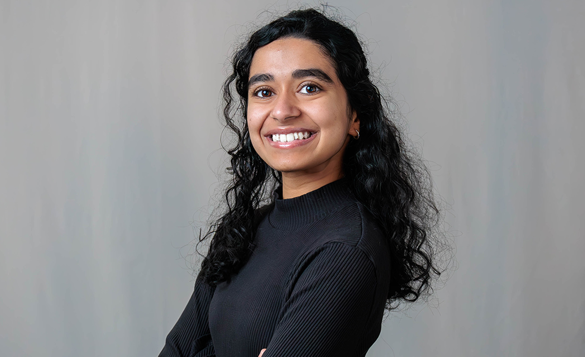 Aarya Newasekar poses smiling, arms crossed, wearing a black sweater and standing in front of a grey backdrop.
