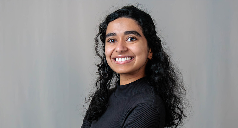 Aarya Newasekar poses smiling, arms crossed, wearing a black sweater and standing in front of a grey backdrop.