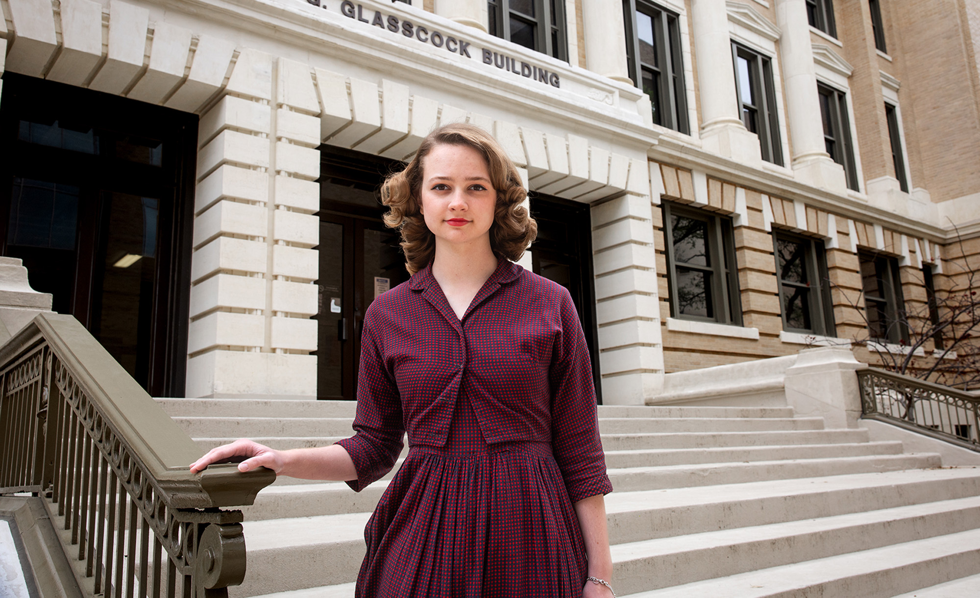 An individual standing confidently on the steps in front of the Melbern G. Glasscock Building, wearing a burgundy dress and a serious expression.