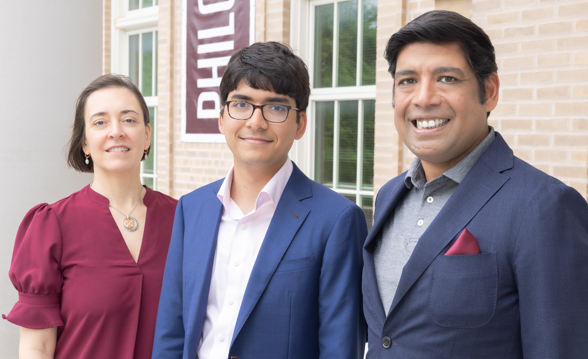 Three people standing together in front of a brick building, smiling at the camera. They are dressed in business attire.