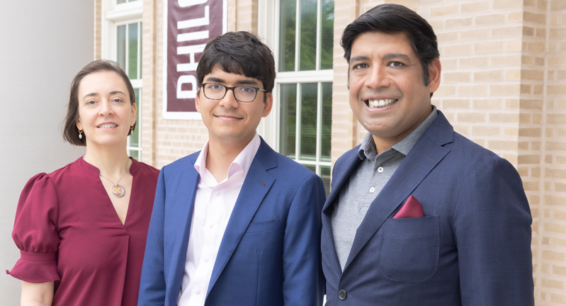 Three people standing together in front of a brick building, smiling at the camera. They are dressed in business attire.