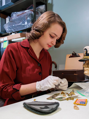 A museum curator examining artifacts at a desk, wearing gloves and surrounded by historical objects.