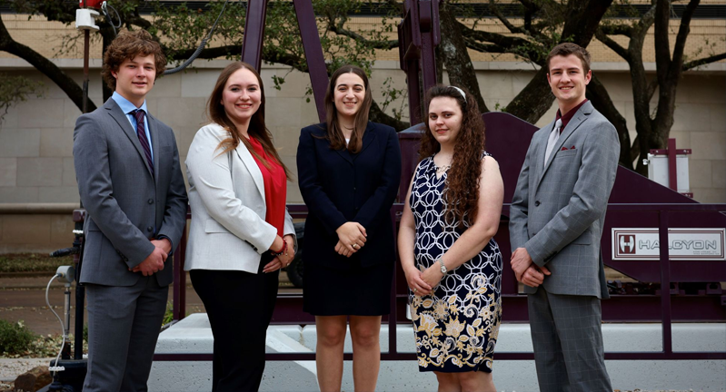 Five students dressed in business attire standing in front of a large maroon structure, smiling at the camera.